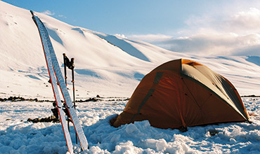 In einer verschneiten Landschaft mit schneebedeckten Bergen im Hintergrund steht ein Zelt. Unter einem klaren, blauen Himmel lehnen zwei Skier und Stöcke am Zelt.