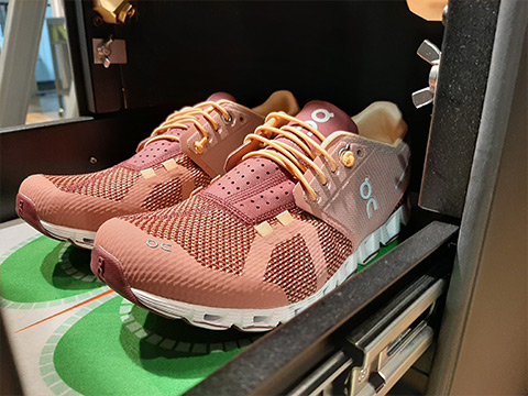 Auf einem Regal steht ein Paar rosa-orangefarbene Sportschuhe. Sie haben ein Netzdesign mit kontrastierenden weißen und grünen Sohlen. Die Schnürsenkel sind gelb-orange und an der Seite ist ein kleines „oc“-Logo zu sehen.