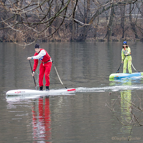 Zwei Personen fahren Stand-Up-Paddleboard auf einem ruhigen Fluss. Die Person vorne trägt ein rotes Outfit und einen roten Hut, während die Person dahinter ein hellgrünes Outfit und einen Helm trägt. Im Hintergrund säumen kahle Bäume das Flussufer.