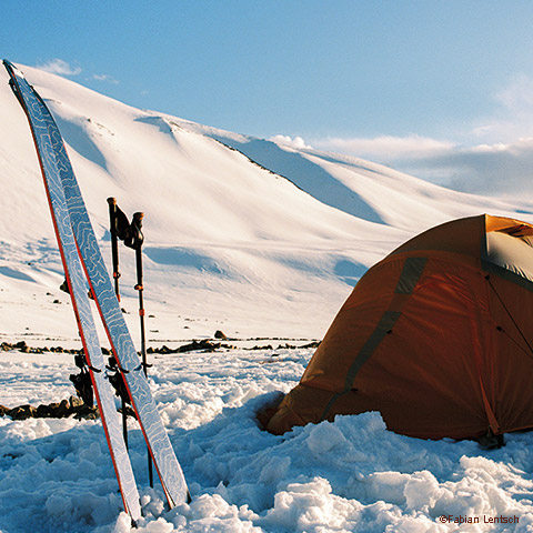 Ein leuchtend orangefarbenes Zelt steht in einer verschneiten Landschaft, in der ein Paar Skier und Stöcke aufrecht stehen. Im Hintergrund ist unter einem klaren blauen Himmel ein großer, schneebedeckter Berg zu sehen.