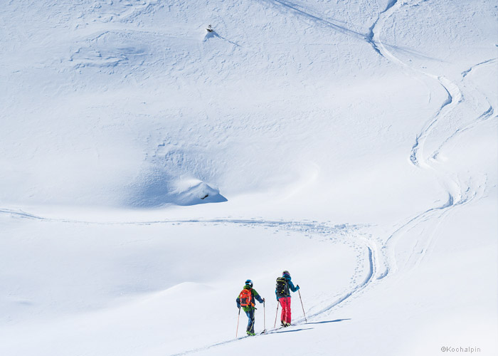 Zwei Skifahrer in heller Ausrüstung laufen unter einem bewölkten Himmel einen weiten, schneebedeckten Hang hinauf. Spuren von früheren Skifahrern durchziehen den Schnee und führen hinauf zu einem Hügel, auf dem in der Ferne eine einsame Person zu sehen ist.