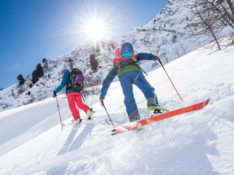 Zwei Menschen in bunter Skiausrüstung erklimmen bei strahlendem Sonnenschein einen schneebedeckten Berg. Sie stützen sich mit Skistöcken ab und tragen Rucksäcke auf dem Rücken, umgeben von einem klaren blauen Himmel und einer schneebedeckten Landschaft.