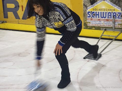 Person in einem gemusterten Pullover spielt Eisstockschießen auf einer Eisbahn und beugt sich nach vorne, um die Stange schieben zu lassen. Im Hintergrund sind gelbe Werbetafeln zu sehen.