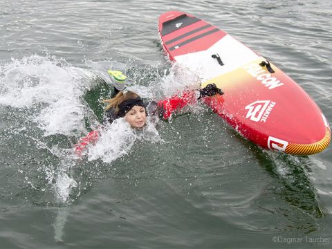 Person in roter Jacke fällt neben einem umgekippten roten Paddleboard ins Wasser. Sie hat einen Fuß in der Luft und das Wasser spritzt herum.