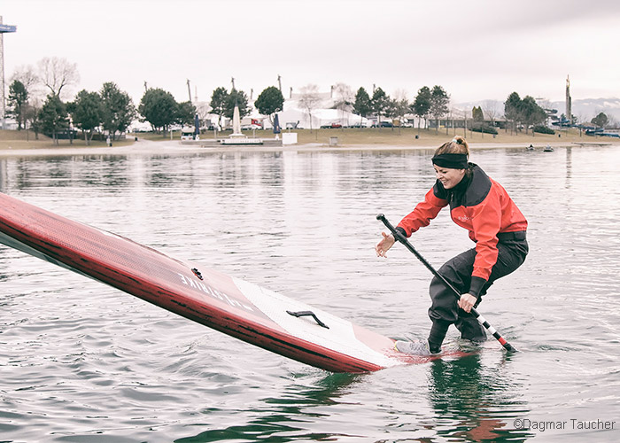 Eine Person in einer roten Jacke und einem schwarzen Stirnband balanciert mit einem Paddel in der Hand auf einem Stand-Up-Paddleboard auf einem ruhigen Gewässer. Im Hintergrund sind Bäume und eine parkähnliche Fläche unter einem bewölkten Himmel zu sehen.