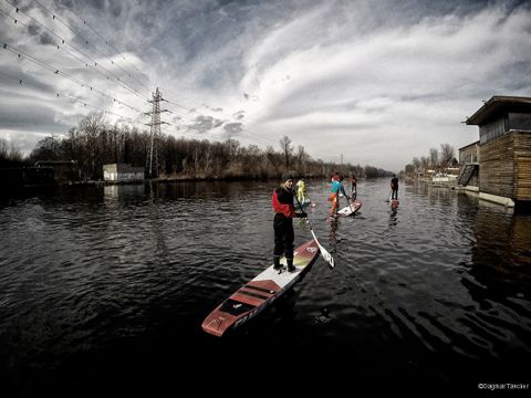 Vier Personen paddeln auf einem ruhigen Wasserweg unter einem bewölkten Himmel. Im Hintergrund säumen Bäume und Stromleitungen das Ufer, rechts ist ein Holzgebäude zu sehen.