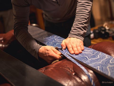Eine Person im Langarmhemd schärft mit einer Feile und festem Druck die Kanten eines Skis. Der Ski weist ein blaues, welliges Muster auf. Sie arbeiten auf einer braunen Lederoberfläche in einer warm beleuchteten Umgebung.