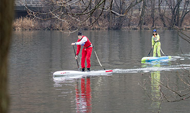Zwei Personen fahren auf einem ruhigen Gewässer Stand-Up-Paddleboard. Einer trägt ein rotes Outfit, der andere ein gelbes. Im Hintergrund sind kahle Bäume und eine Brücke zu sehen, die einen kühlen, bewölkten Tag widerspiegeln.