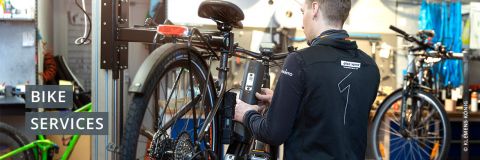 Eine Person repariert in einer Werkstatt ein Fahrrad, das auf einem Ständer montiert ist. Auf der Rückseite ihres Hemdes ist eine große Zahl „1“ aufgedruckt. Im Hintergrund sind verschiedene Werkzeuge und Fahrradteile zu sehen. Der Text auf der linken Seite lautet „Fahrradservice“.