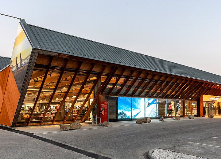 Ein modernes Gebäude mit einer einzigartigen Schrägdachkonstruktion mit großen Glasfenstern und Holzelementen vor einem klaren Himmel. In der oberen rechten Ecke befindet sich ein „German Design Award Winner 2020“-Schild. Das Äußere ist warm beleuchtet.