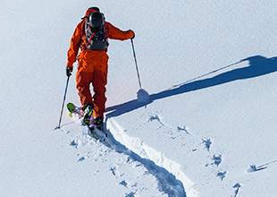 Ein Skifahrer in orangefarbener Ausrüstung und mit Rucksack fährt einen schneebedeckten Hang entlang und hinterlässt dabei eine schmale Spur. Der klare blaue Himmel wirft einen deutlichen Schatten auf den hellen Schnee.
