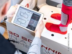 Eine Person hält ein Tablet mit Schuhanpassungsdaten neben den Füßen einer Person, die rote und graue Socken trägt. Sie steht auf einer Plattform für einen 3D-Fußscan. Im Hintergrund ist ein Teil einer Maschine mit der Aufschrift „3D-SCANNEN“ zu sehen.