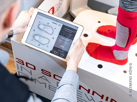 Eine Person hält ein Tablet mit Schuhanpassungsdaten neben den Füßen einer Person, die rote und graue Socken trägt. Sie steht auf einer Plattform für einen 3D-Fußscan. Im Hintergrund ist ein Teil einer Maschine mit der Aufschrift „3D-SCANNEN“ zu sehen.