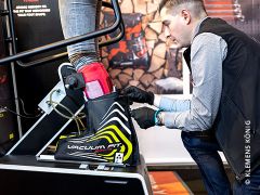 Eine Person passt mithilfe einer Vakuum-Anpassmaschine einen Skischuh für einen Kunden an. Der Kunde steht auf einer Plattform und trägt den Schuh, während der Techniker die Einstellungen vornimmt. Um ihn herum sind verschiedene Geräte und Informationsdisplays.
