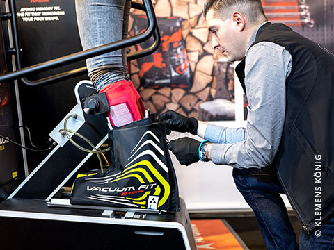 Eine Person passt mithilfe einer Vakuum-Anpassmaschine einen Skischuh für einen Kunden an. Der Kunde steht auf einer Plattform und trägt den Schuh, während der Techniker die Einstellungen vornimmt. Um ihn herum sind verschiedene Geräte und Informationsdisplays.