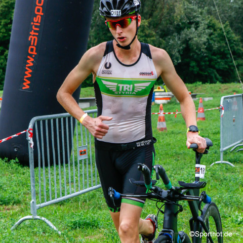 Ein Athlet in einem schwarz-grünen Triathlonanzug und mit Sonnenbrille läuft während eines Triathlons mit einem Fahrrad in der Hand. Im Hintergrund sind Bäume, ein großer aufblasbarer Bogen und Metallbarrikaden zu sehen.