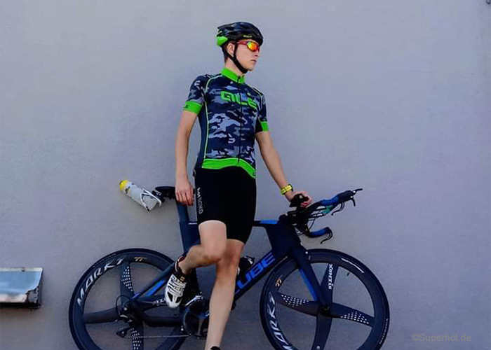 Eine Person in Radsportbekleidung, inklusive Helm und Sonnenbrille, steht mit einem Fuß auf einem blauen Rennrad. Der Radfahrer lehnt an einer grauen Wand und blickt zur Seite. Am Fahrradrahmen ist eine Wasserflasche befestigt.