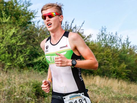 Sportler mit Sonnenbrille und ärmellosem Triathlonanzug läuft im Freien auf einer Grasbahn. Er trägt eine schwarze Uhr und seine Startnummer ist 481. Im Hintergrund sind grüne Bäume und ein bewölkter blauer Himmel zu sehen.