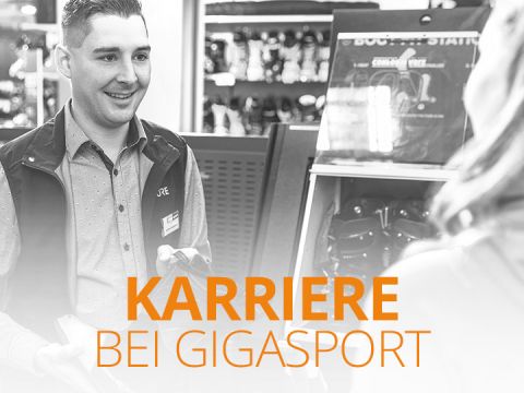 Ein Verkäufer lächelt, während er einen Kunden in einem Geschäft bedient. Der Text „KARRIERE BEI GIGASPORT“ ist in Orange hervorgehoben. Im Hintergrund des Geschäfts stehen verschiedene Produkte in Regalen.
