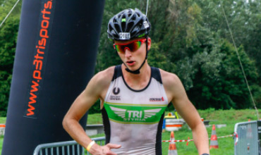 Eine Person im Triathlon-Outfit und mit Helm läuft an einem großen aufblasbaren Bogen mit der Aufschrift „p3trisports“ vorbei. Sie befinden sich in einer Außenumgebung, im Hintergrund sind Bäume und orangefarbene Kegel zu sehen.