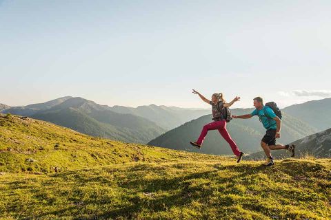 Zwei Wanderer mit Rucksäcken springen fröhlich durch eine Berglandschaft. Die Szene ist sonnig, mit sanften grünen Hügeln und fernen Bergen unter einem klaren blauen Himmel. Ein Wanderer streckt die Hand nach dem anderen aus und fängt einen Moment der Aufregung und des Abenteuers ein.