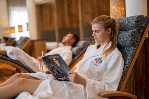 Eine Frau im weißen Bademantel liest eine Modezeitschrift, während sie in einem gepolsterten Sessel in einem Spa liegt. Im Hintergrund entspannt sich ein Mann, ebenfalls im Bademantel. Die Umgebung ist ruhig und luxuriös, mit Holzdekor und sanfter Beleuchtung.