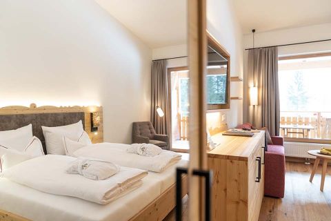 Ein gemütliches Hotelzimmer mit einem großen Bett, das mit weißer Bettwäsche und Handtüchern ausgestattet ist. Es gibt eine Holzkommode, einen an der Wand montierten Fernseher und einen Balkon mit Aussicht. Sanfte Beleuchtung und Vorhänge vervollständigen die warme, einladende Atmosphäre.