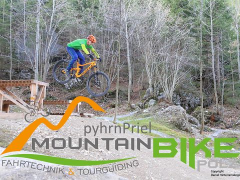 Ein Radfahrer in grüner Jacke und blauer Hose springt mit einem Mountainbike über einen von Bäumen umgebenen Feldweg. Im Vordergrund steht der Text: „PyhrnPriel Mountain Bike Fahrtechnik & Tourguiding.“.