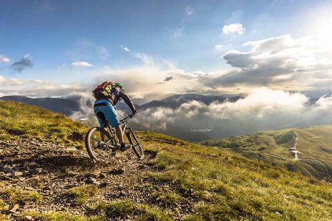 Ein Radfahrer in blauen Shorts und mit Rucksack fährt einen felsigen Bergpfad hinunter. Die Szene ist voller grüner Hügel unter einem teilweise bewölkten Himmel, wobei Nebel und Sonnenlicht eine dramatische Atmosphäre schaffen.