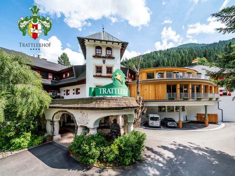 Vorderansicht des Hotels Trattlerhof in einer Bergregion mit blauem Himmel. Das Gebäude zeichnet sich durch eine Mischung aus traditioneller und moderner Architektur aus. Es gibt viel Grün und im Hintergrund Hügel. Das Logo des Hotels ist in der oberen linken Ecke sichtbar.