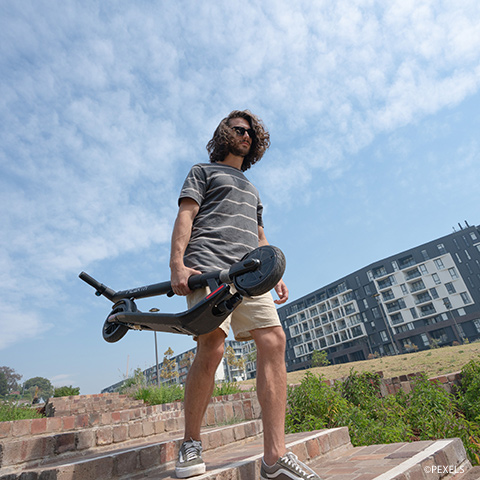 Eine Person mit lockigem Haar und Sonnenbrille trägt einen Elektroroller und steht auf Ziegelstufen. Sie trägt ein gestreiftes Hemd und Shorts. Im Hintergrund sind moderne Gebäude und ein blauer Himmel mit vereinzelten Wolken zu sehen.
