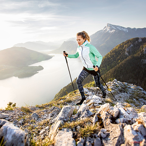 Eine Frau wandert mit Trekkingstöcken auf einem felsigen Bergpfad. Sie trägt eine hellgrüne und weiße Jacke und gemusterte Leggings. Im Hintergrund ist eine malerische Aussicht auf einen See und Berge in der Ferne unter einem klaren Himmel zu sehen.