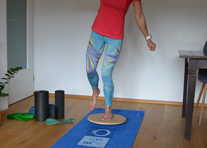 Eine Person in einem roten Hemd und bunten Leggings balanciert auf einem hölzernen Balance Board, das auf einer blauen Trainingsmatte steht. In der Nähe liegen Schaumstoffrollen und Widerstandsbänder auf einem Holzboden.