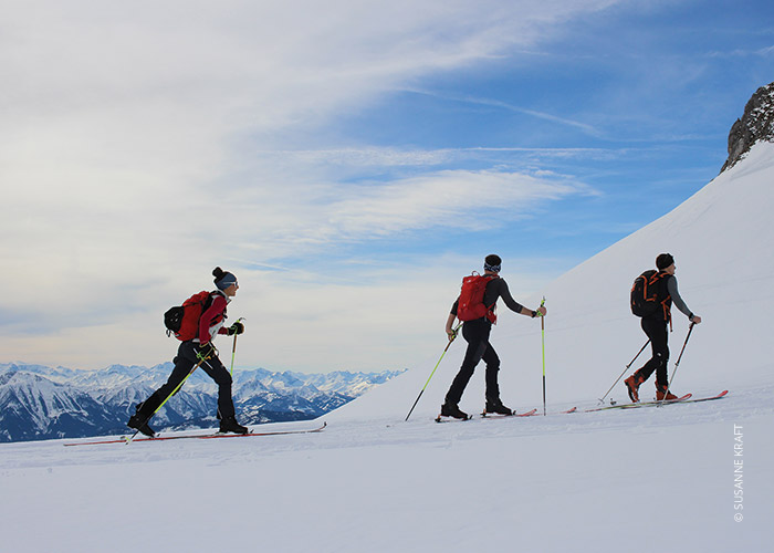 Drei Personen in Winterkleidung und Rucksäcken fahren einen schneebedeckten Berg hinauf. Der Himmel ist teilweise bewölkt und im Hintergrund ist eine Bergkette zu sehen.