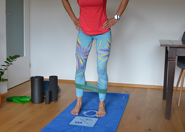 Eine Person in einem roten Hemd und gemusterten Leggings steht auf einer blauen Trainingsmatte und hat ein Widerstandsband um ihre Waden. In der Nähe befinden sich zwei Schaumstoffrollen und rechts ist teilweise ein Tisch sichtbar.