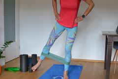 Person, die zu Hause trainiert, trägt ein rotes Hemd und gemusterte Leggings. Sie verwendet ein Widerstandsband um ihre Knöchel für Beinübungen. Im Raum sind Yogamatte, Schaumstoffrollen und eine Pflanze zu sehen.