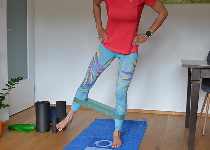 Person, die zu Hause trainiert, trägt ein rotes Hemd und gemusterte Leggings. Sie verwendet ein Widerstandsband um ihre Knöchel für Beinübungen. Im Raum sind Yogamatte, Schaumstoffrollen und eine Pflanze zu sehen.
