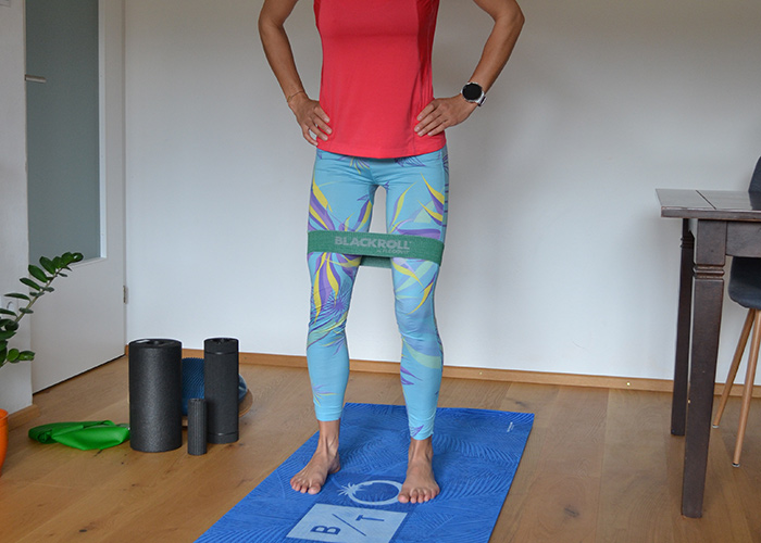 Eine Person steht auf einer blauen Trainingsmatte. Sie trägt ein rotes Oberteil und bunte Leggings und hat ein Widerstandsband über den Knien. Im Hintergrund sind drei Schaumstoffrollen neben einer Pflanze und einem Holztisch zu sehen.