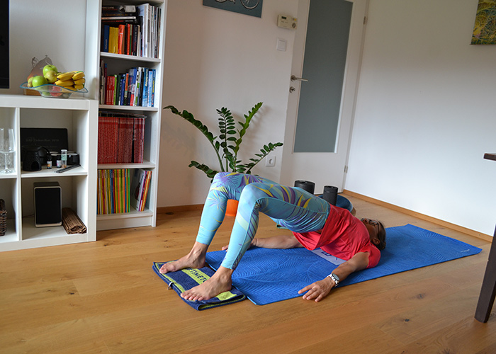 Person, die eine Bridge-Yoga-Pose auf einer blauen Matte in einem Wohnzimmer ausführt. Sie trägt ein rotes Hemd und gemusterte Leggings. Im Hintergrund gibt es ein Bücherregal mit Büchern und Früchten, eine Pflanze und eine geschlossene Tür.