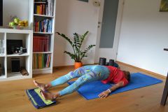 Eine Person in einem roten Hemd und gemusterten Leggings liegt auf einer blauen Yogamatte in einem Wohnzimmer. Der Raum hat einen Holzboden, ein Bücherregal mit verschiedenen Gegenständen, eine Topfpflanze und eine Obstschale. Sie scheint sich zu entspannen oder zu meditieren.