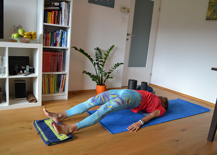 Eine Person in einem roten Hemd und gemusterten Leggings liegt auf einer blauen Yogamatte in einem Wohnzimmer. Der Raum hat einen Holzboden, ein Bücherregal mit verschiedenen Gegenständen, eine Topfpflanze und eine Obstschale. Sie scheint sich zu entspannen oder zu meditieren.