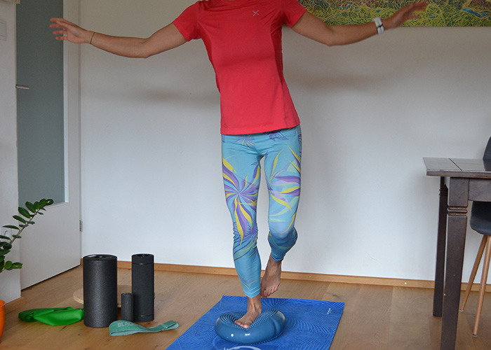 Eine Person balanciert auf einem blauen Trainingskissen auf einer Yogamatte. Sie trägt ein rotes Hemd und bunte Leggings. Trainingsgeräte wie Schaumstoffrollen und Widerstandsbänder liegen in der Nähe auf dem Holzboden.
