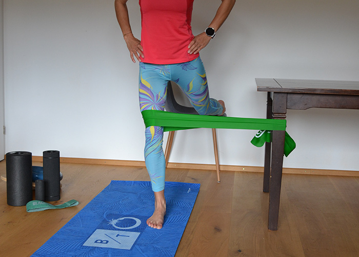 Eine Person in einem roten Hemd und bunten Leggings führt mithilfe eines grünen Widerstandsbands, das an einem Tisch befestigt ist, eine Beinstreckübung durch. Sie steht auf einer blauen Trainingsmatte mit Schaumstoffrollen in der Nähe.