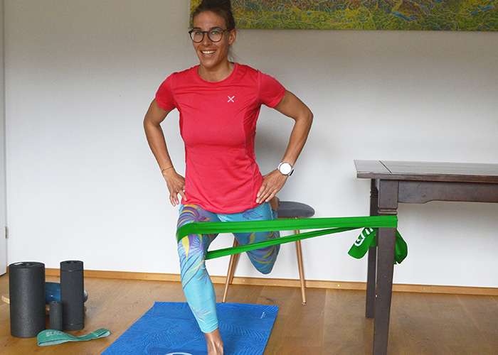 Eine Person in einem roten Hemd und blauen Leggings trainiert mit einem grünen Widerstandsband unter ihrem Knie, das von einem Tisch gestützt wird. Sie lächelt und steht auf einer blauen Matte mit Schaumstoffrollen in der Nähe.
