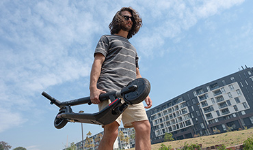 Eine Person mit langen Haaren und Sonnenbrille trägt einen Elektroroller im Freien. Sie trägt ein gestreiftes Hemd und Shorts. Im Hintergrund sind moderne Wohnhäuser unter einem teilweise bewölkten Himmel zu sehen.