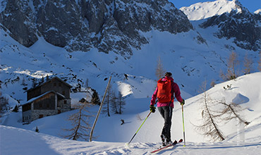 Eine Person mit rotem Rucksack und Skiausrüstung fährt auf einem schneebedeckten Berghang Ski. In der Ferne ist eine Hütte zu sehen. Im Hintergrund sind hoch aufragende, felsige Berge und ein klarer blauer Himmel zu sehen. Vereinzelte Bäume sind über die Landschaft verstreut.