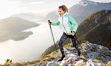 Eine Person wandert mit Trekkingstöcken einen felsigen Bergpfad hinauf. Sie trägt eine grün-weiße Jacke und gemusterte Leggings. Der Hintergrund zeigt eine malerische Aussicht auf einen See und entfernte Berge unter einem klaren Himmel.