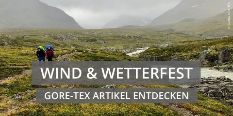 Zwei Wanderer mit Rucksäcken gehen einen schmalen Pfad durch eine Berglandschaft entlang. Der Himmel ist bedeckt, im Hintergrund ist eine Brücke zu sehen. Das Bild zeigt eine Textüberlagerung, die für Gore-Tex-Produkte wirbt.