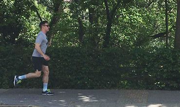 Eine Person joggt auf einem gepflasterten Weg, umgeben von üppigen grünen Bäumen und Büschen. Sie trägt ein graues Hemd, schwarze Shorts, blau-weiß gestreifte Socken, Turnschuhe und eine Sonnenbrille. Die Szene ist sonnig und im Freien.