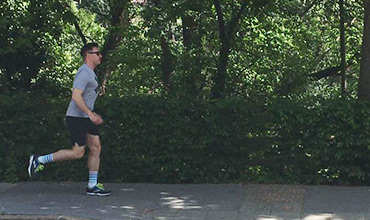 Eine Person joggt auf einem gepflasterten Weg, umgeben von üppigen grünen Bäumen und Büschen. Sie trägt ein graues Hemd, schwarze Shorts, blau-weiß gestreifte Socken, Turnschuhe und eine Sonnenbrille. Die Szene ist sonnig und im Freien.